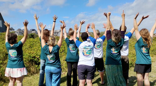 Gruppenbild, 8 Personen von hinten. Alle tragen ein "Surf and Heal" T-Shirt und strecken die Arme nach oben. Sie stehen auf einer Wiese.